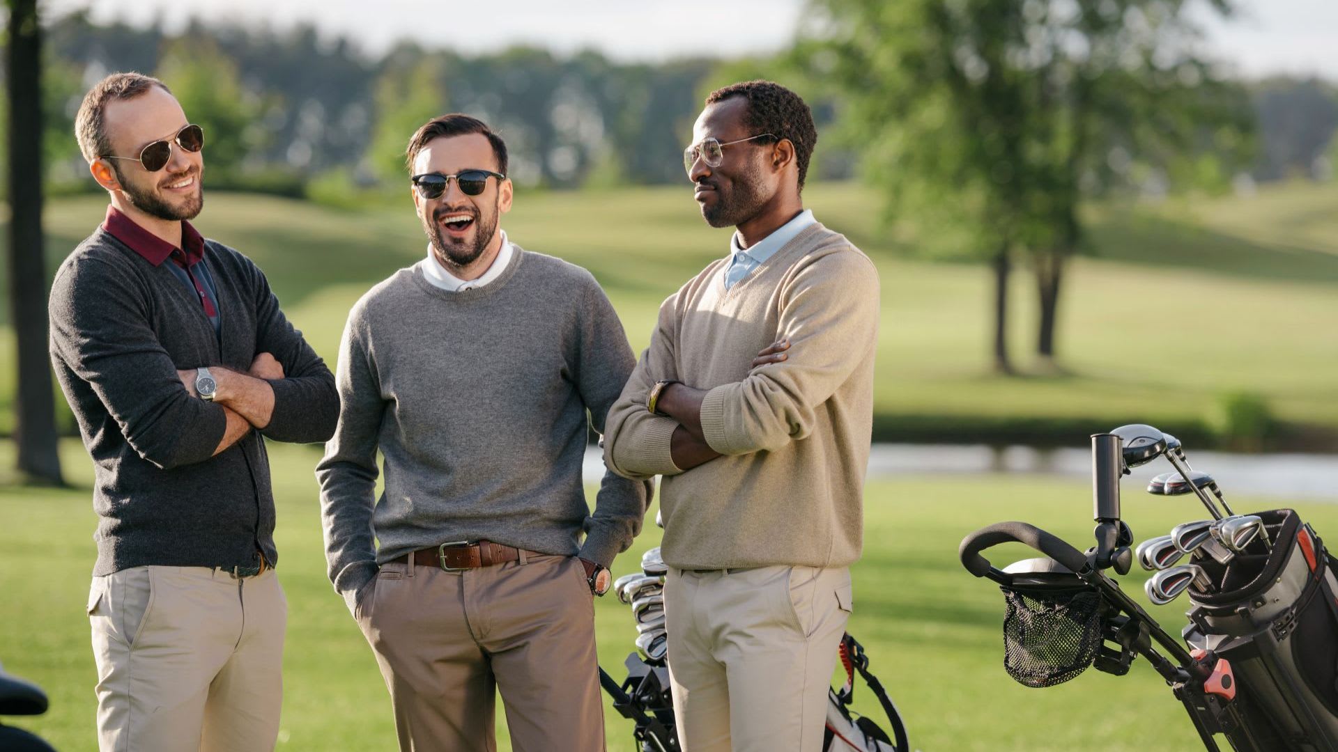 Three men in sweaters and sunglasses stand on a golf course, laughing and talking, with golf bags from top golf brands and clubs visible in the foreground.